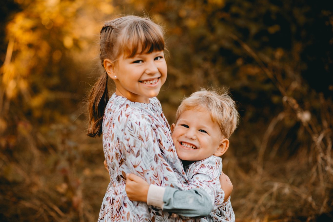 Zwei lachende Kinder umarmen sich in einer herbstlichen Landschaft.