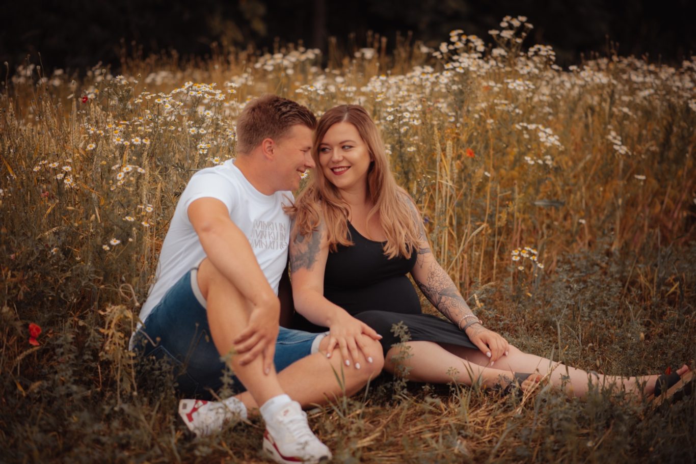 Ein Paar sitzt in einer blühenden Wiese und lächelt sich glücklich an.