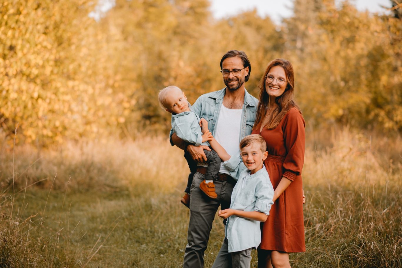 Familie steht in einem herbstlichen Feld, der Vater hält ein Kind, die Mutter lächelt.